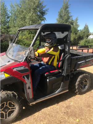 Red 4-wheel ATV with black canopy. Driver in blue jeans, yellow and brown shirt, black helmet.