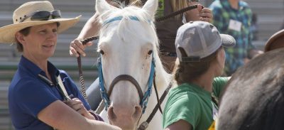 Person in blue shirt and straw hat watching youth ride a white horse.