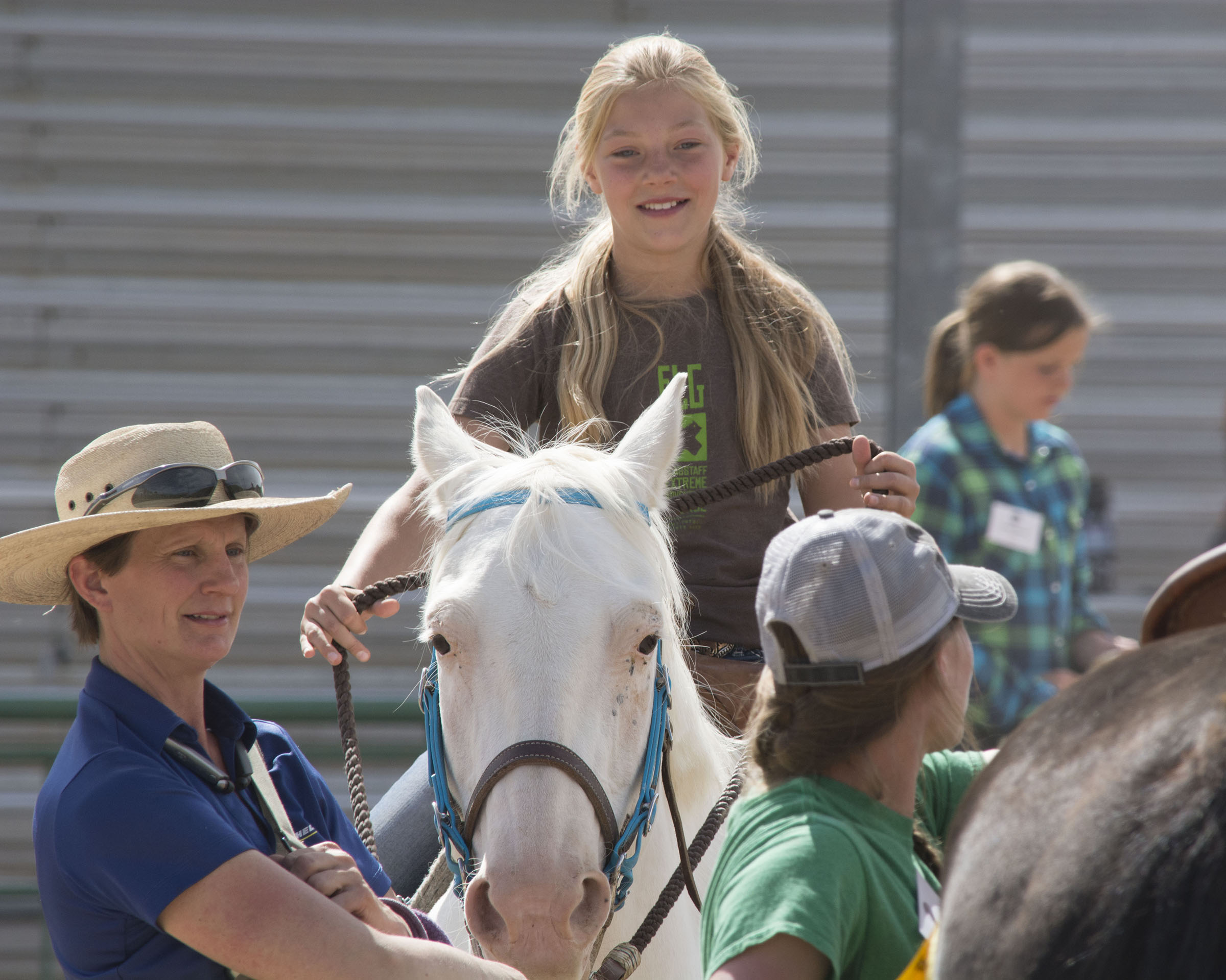 Woman in blue shirt and straw hat watching while youth rides white horse.