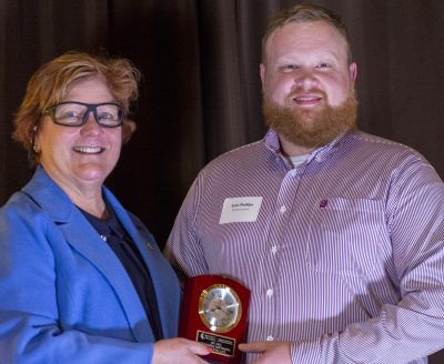 woman in blue shirt and glasses presents bearded man with an award plaque