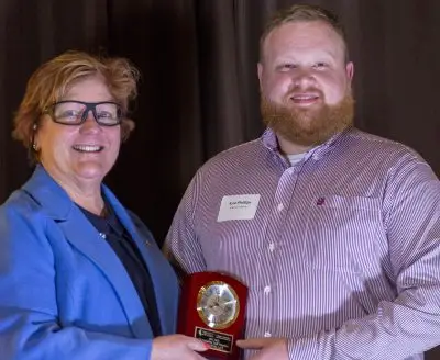 woman in blue shirt and glasses presents bearded man with an award plaque