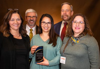 Group photo with a woman in a turquoise shirt holding an award plaque and surrounded by four other people