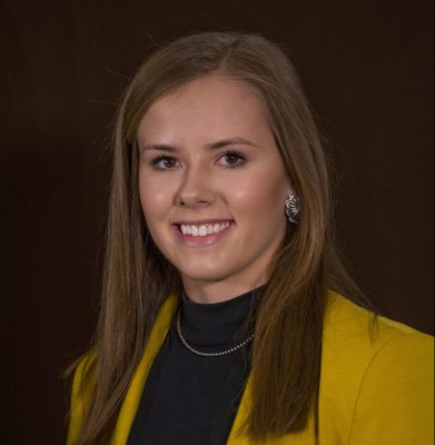 headshot of smiling woman with brown hair wearing black shirt and yellow jacket