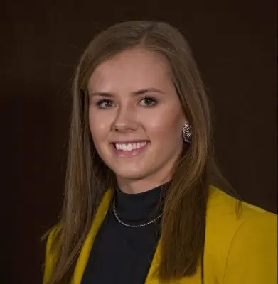 headshot of smiling woman with brown hair wearing black shirt and yellow jacket