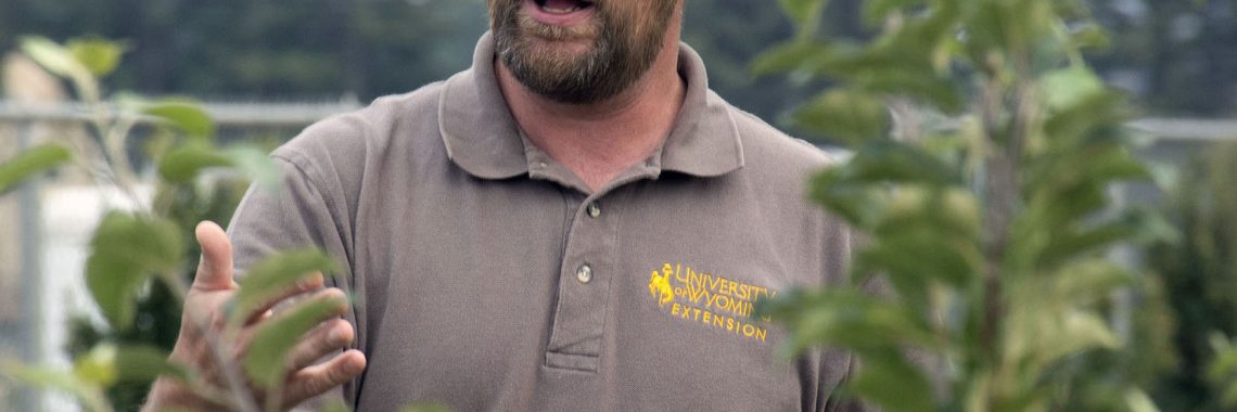 man wearing brown and yellow University of Wyoming baseball cap and polo shirt stands next to a tree and talks