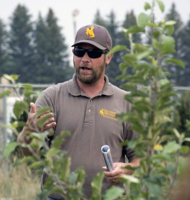 man wearing brown and yellow University of Wyoming baseball cap and polo shirt stands next to a tree and talks