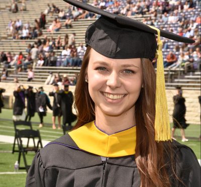 headshot of woman wearing black and yellow graduation hat and robe stands in a stadium