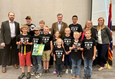 eleven students wearing black t-shirts with Laramie County 4-H Robotics logo stand with four adults beside a flag
