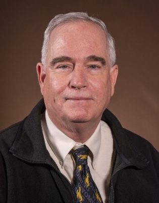 portrait of smiling man wearing vest over white shirt and brown tie with gold cowboy logo