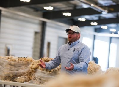 Bearded man wearing glasses and baseball cap stands in front of bags of wool