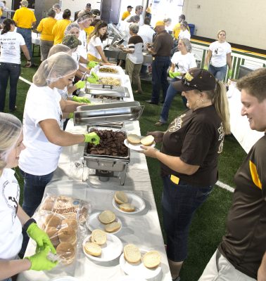 students wearing white t-shirts and hairnets serve food to people wearing brown and gold attire