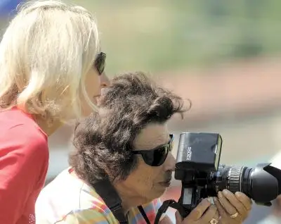 two women wearing sunglasses stand together, one holding a camera