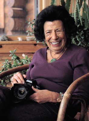 smiling woman wearing purple shirt seated and holding a camera