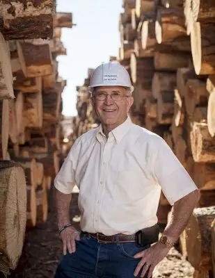smiling man wearing white hardhat and standing between two piles of logs