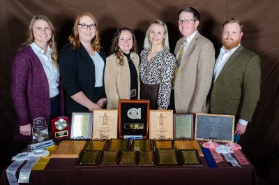 four women and two men stand behind a table covered in award plaques