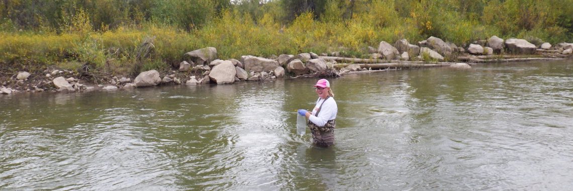 woman in baseball cap and waders collects water sample in river