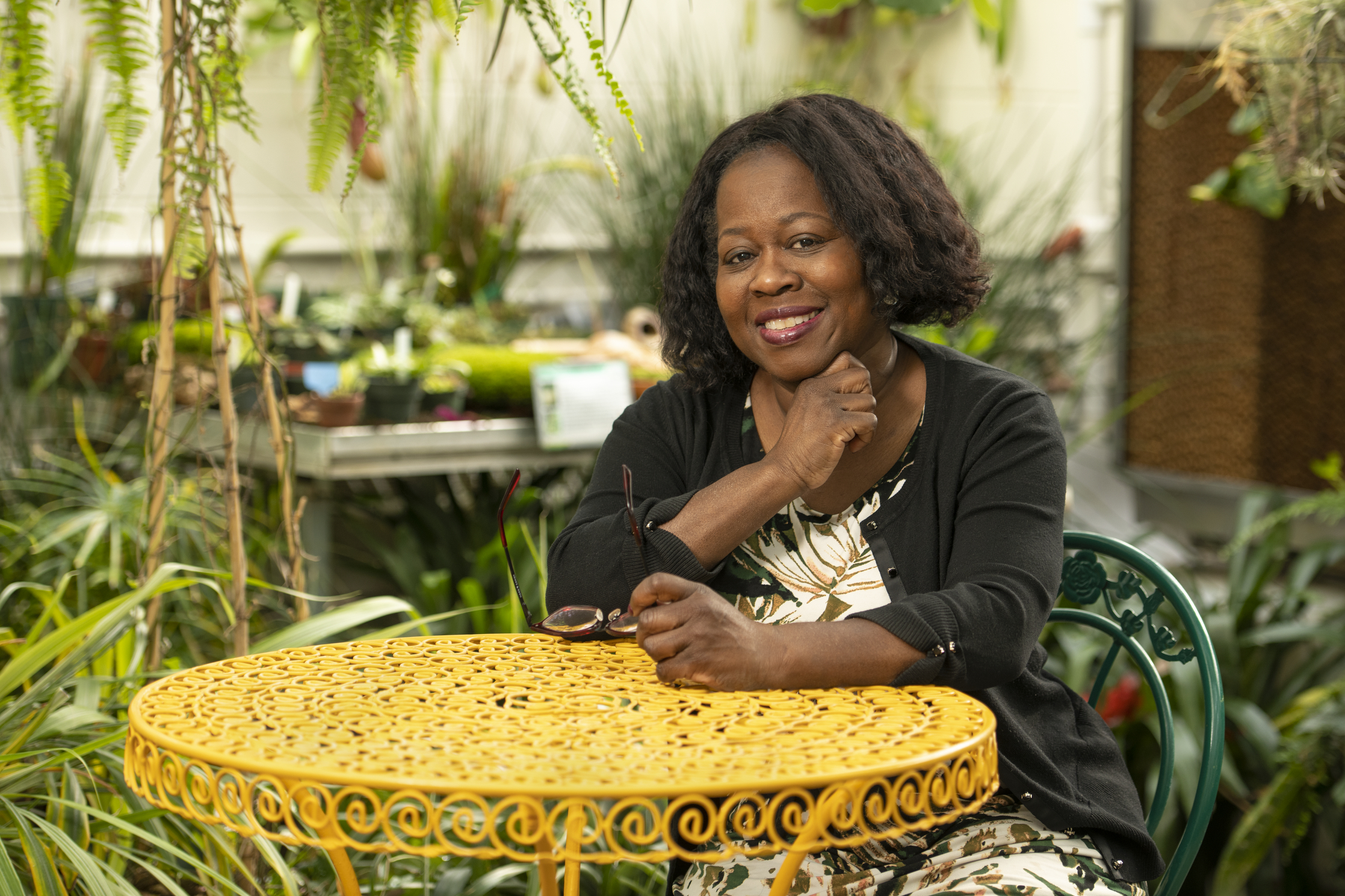 smiling woman wearing a black sweater and flowered dress sits at a yellow table surrounded by plants