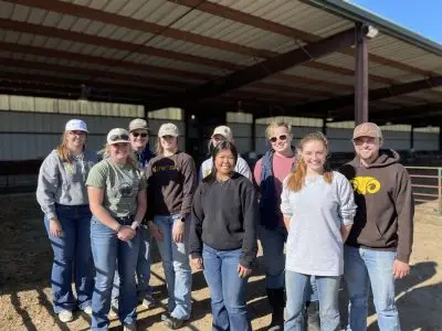Group photo of nine students, all wearing jeans, standing in front of a barn