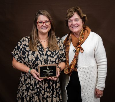 smiling woman wearing red-rimmed glasses and dress with brown and tan pattern holds an award plaque while standing next to a smiling woman in a white sweater and patterned scarf