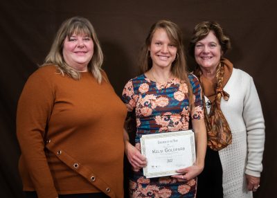 three smiling women stand together, with the one in the middle holding an award plaque