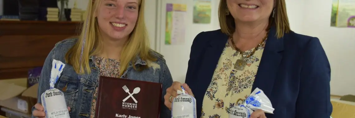 smiling girl holding package of sausage and award plaque stands next to smiling woman wearing blue blazer and holding two packages of sausage