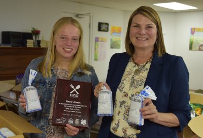 smiling girl holding package of sausage and award plaque stands next to smiling woman wearing blue blazer and holding two packages of sausage