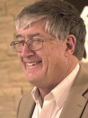 smiling man with grey hearing wearing glasses and a tan coat