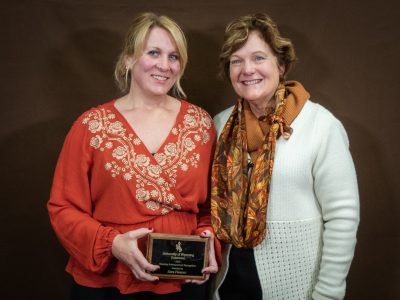 smiling woman with blond hair and reddish shirt holds award plaque and stands next to smiling woman in white sweater and patterned scarf