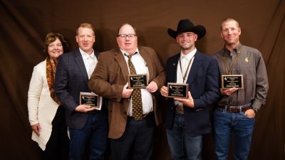 woman wearing white sweater and patterned scarf stands with a group of four men wearing jeans and collared shirts or blazers. All four men are holding award plaques.