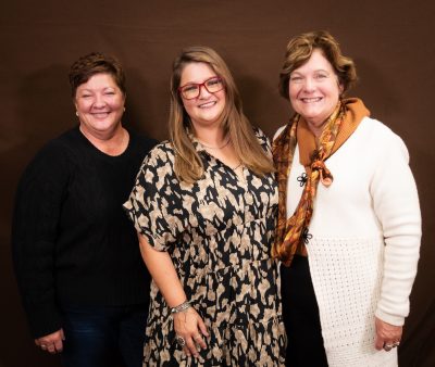 three smiling woman, one wearing black sweater and jeans, one wearing a black and tan dress, and the third wearing a white sweater and orange patterned scarf