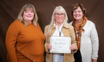 smiling woman in orange sweater and smiling woman in white sweater stand on either side of woman wearing glasses and holding an award plaque