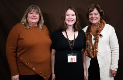 smiling woman in an orange sweater stands beside smiling woman in black dress and smiling woman in white sweater and brown patterned scarf