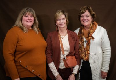 smiling woman wearing burgundy sweater and matching earrings and purse stands between smiling woman in orange sweater and smiling woman in white sweater