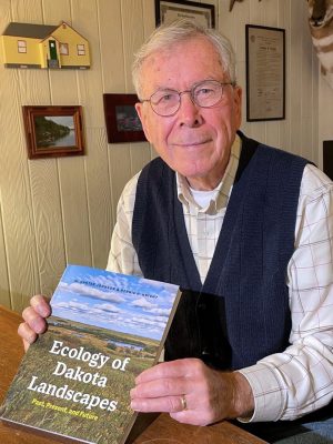 smiling man wearing blue vest and glasses sits at a table holding a book titled Ecology of Dakota Landscapes