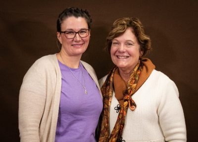 smiling woman wearing purple shirt and off-white sweater stands beside smiling woman in white sweater and patterned orange scarf