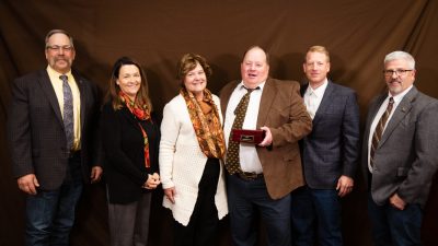 six smiling people pose for a portrait with a man in the center holding a wooden award box.