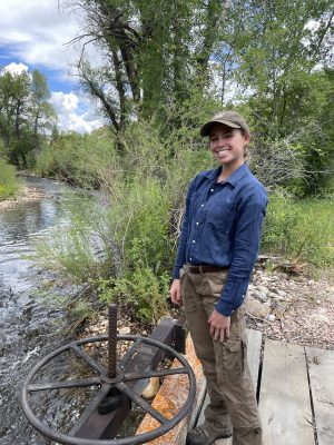 smiling woman wearing baseball cap and cargo pants stands beside a headgate controlling flow in a creek