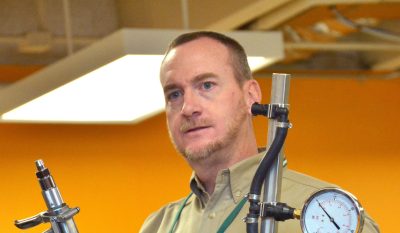 man wearing tan collared shirt and name tag holds up pesticide application equipment during a presentation in a room with orange walls