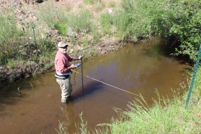 man wearing baseball cap and red shirt stands in the brown water of an irrigation ditch to perform a flow measurement