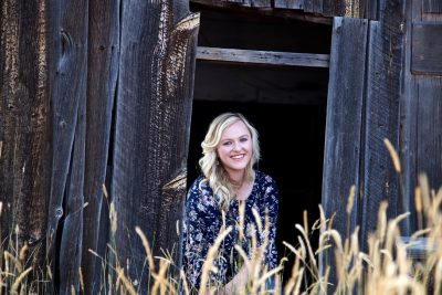 smiling woman with blond hair and blue flowered dress looks out from an old barn into yellow grass
