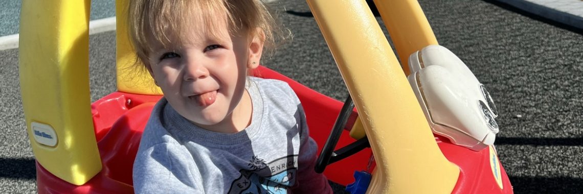 smiling toddler wearing gray shirt and Crocs sticks her tongue out while riding in red and yellow plastic toy car