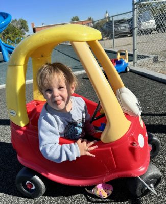 smiling toddler wearing gray shirt and Crocs sticks her tongue out while riding in red and yellow plastic toy car
