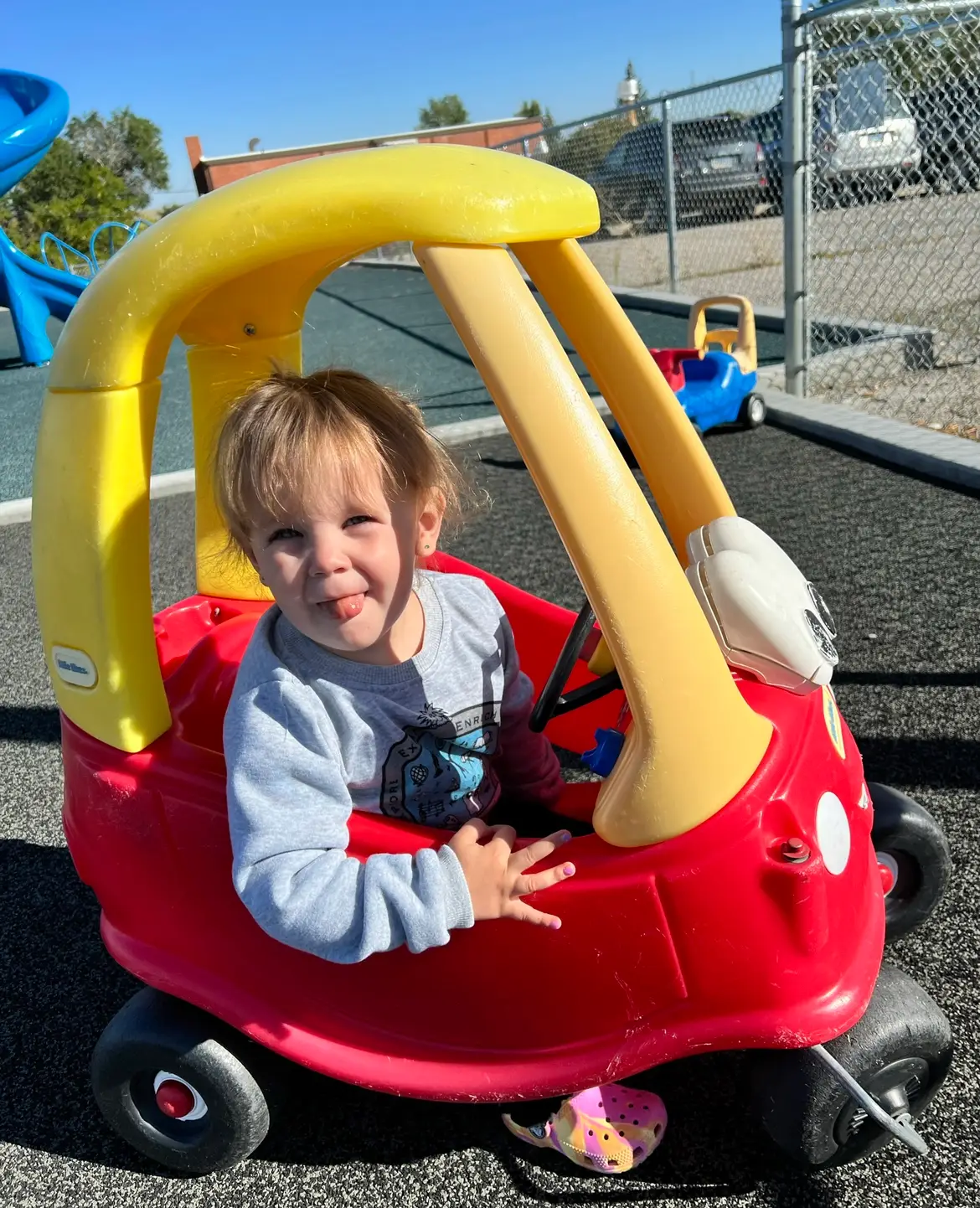 smiling toddler wearing gray shirt and Crocs sticks her tongue out while riding in red and yellow plastic toy car