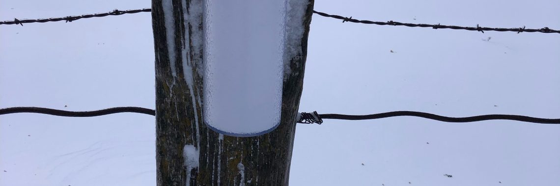 rain gauge filled with snow and attached to a fence post on a barb wire fence