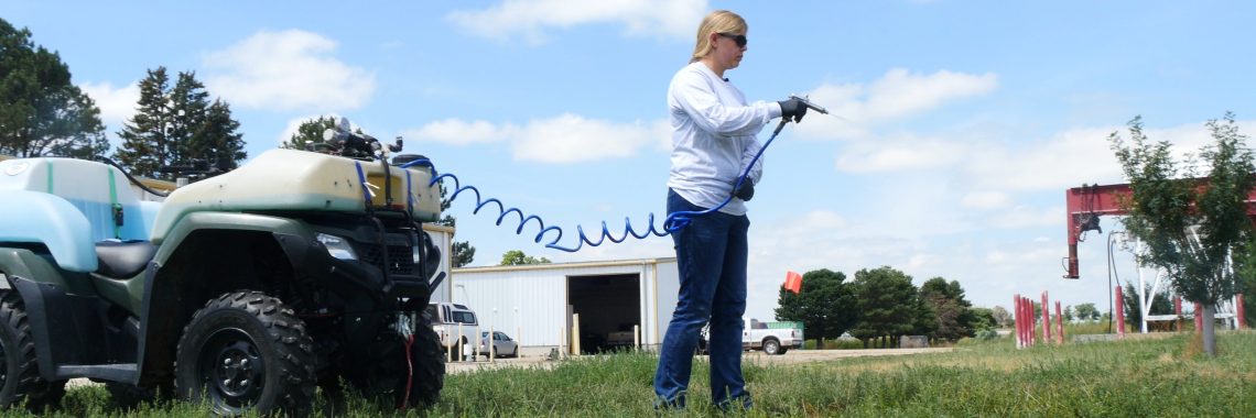 woman wearing long sleeved shirt, jeans, and sunglasses calibrates pesticide sprayer mounted on a four-wheeler