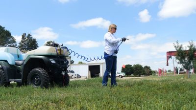 woman wearing long sleeved shirt, jeans, and sunglasses calibrates pesticide sprayer mounted on a four-wheeler