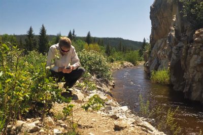 man wearing long-sleeved collared shirt and sunglasses crouches beside an irrigation ditch with water flowing, looking at a phone