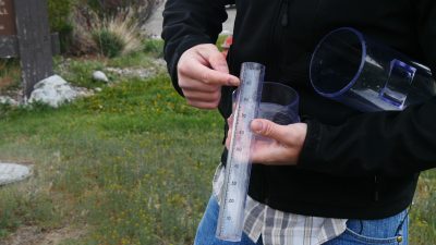 person wearing black sweatshirt points to measurement marks on a rain gauge