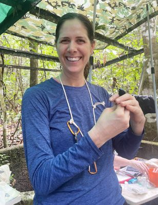 Woman wearing long-sleeved blue shirt holds small blackish blue bird while standing under a structure covered with camouflage fabric