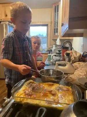 young boy stirs a spoon in a silver bowl next to a glass casserole dish sitting on the stove in the kitchen. An older girl supervises from farther down the counter.